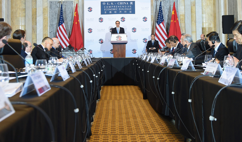 A U.S.–China Comprehensive Economic Dialogue session: delegations seated along a long conference table facing a speaker at a podium, beneath U.S. and Chinese flags and a branded backdrop.